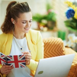 smiling elegant middle age woman in jeans and yellow jacket with laptop taking notes with a pen in a UK flag notebook in the modern house in sunny day.